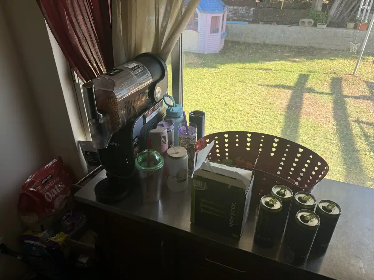 A cluttered kitchen counter next to a window shows a Ninja coffee maker, several colorful water bottles and cups, a bag of…