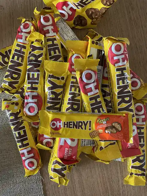 A pile of unwrapped Oh Henry! candy bars with yellow and red wrappers is scattered on a wooden surface, with one Reese's…
