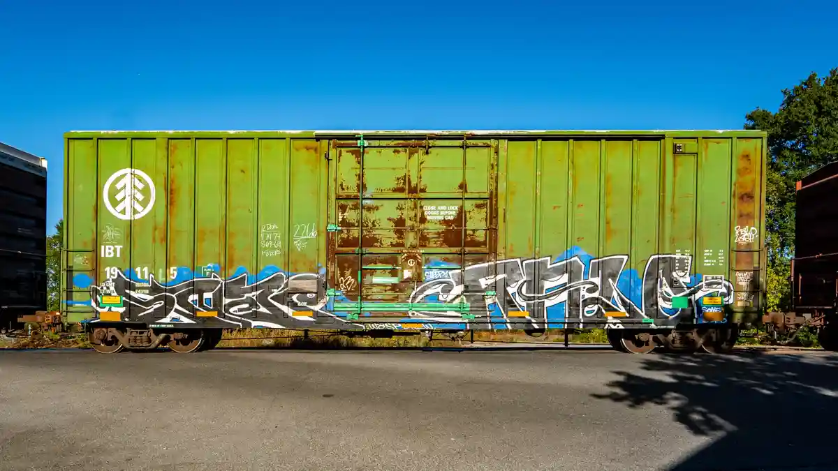 A weathered green freight train car covered in graffiti sits under a clear blue sky, with the text 'CLOSE AND LOCK DOORS…