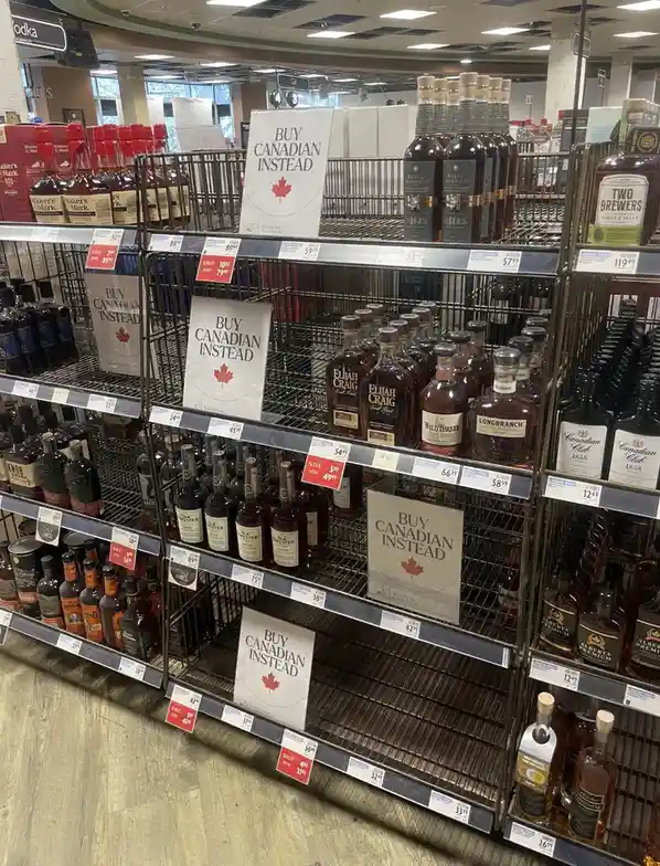 A store aisle filled with liquor shelves, many of which are empty or nearly empty, with multiple white signs that say 'BUY…
