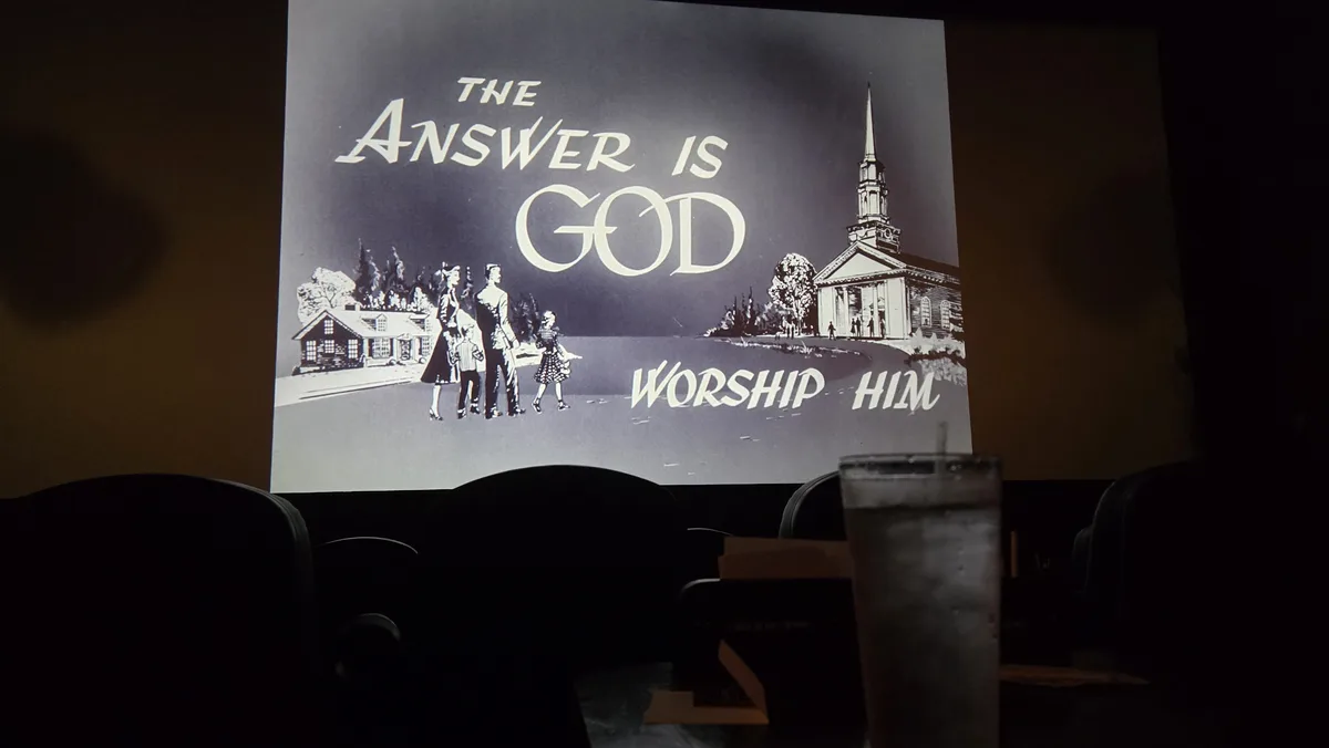 A black-and-white vintage-style illustration is projected onto a screen in a dark theater, showing a family of four walking…