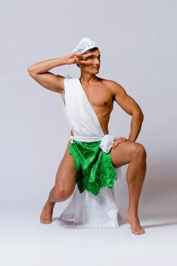 A muscular man in a white toga and green leaf skirt poses in a studio, saluting with one hand while looking off to the side…