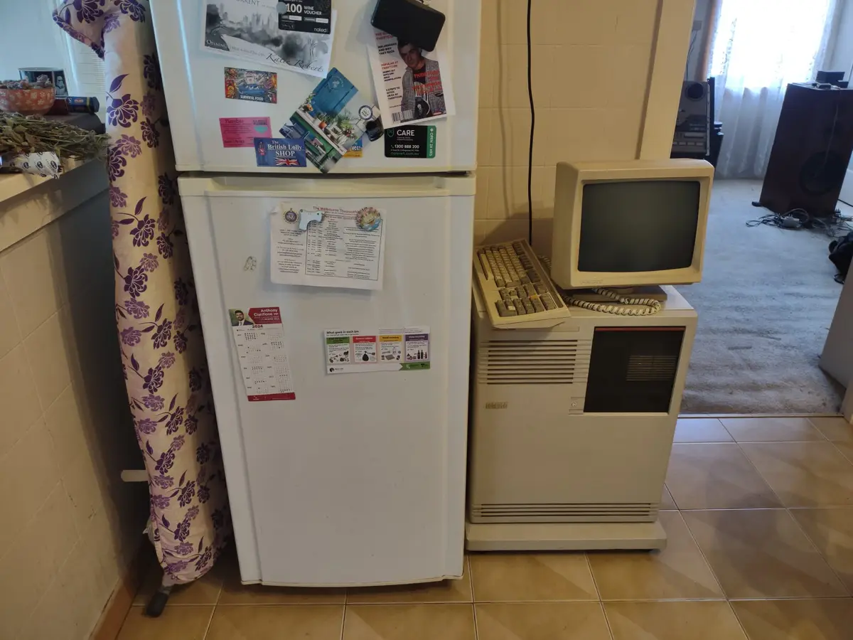 A white refrigerator covered in magnets and papers stands next to a beige, bulky, vintage computer with a CRT monitor and…