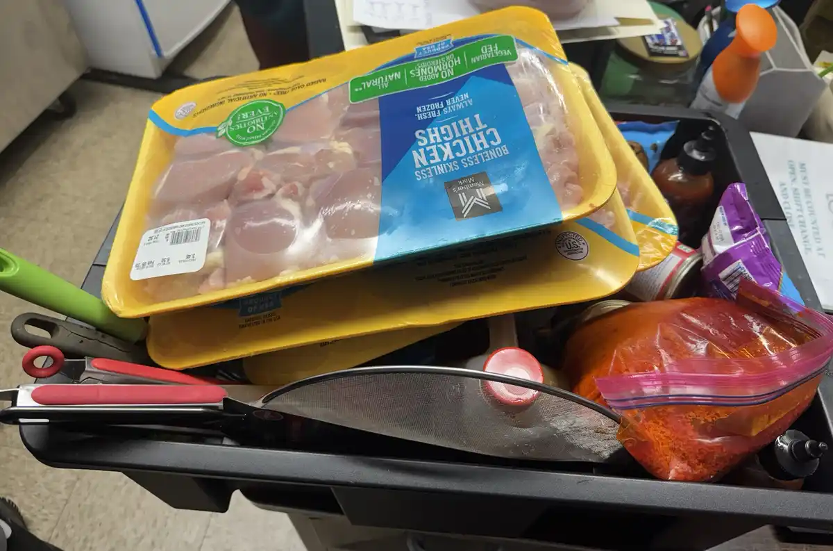 A close-up photo shows two yellow plastic trays of raw boneless skinless chicken thighs stacked on a black kitchen cart…