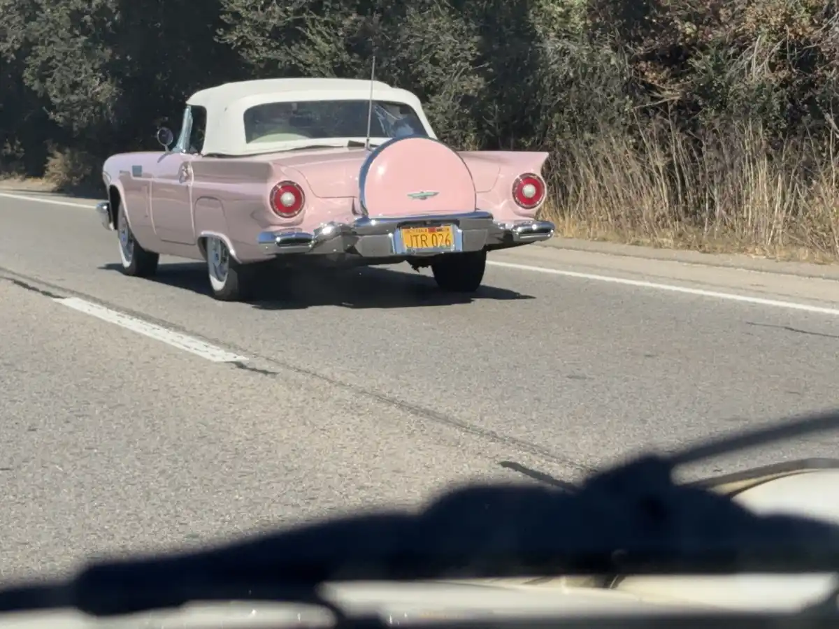 A pink 1950s Ford Thunderbird convertible with a white soft top is driving on a paved road, viewed from behind. The car's…