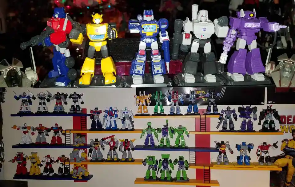 A large collection of Transformers action figures is displayed on multiple shelves, with larger figures at the top and…