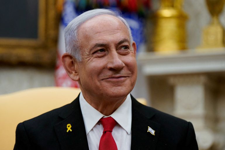 a man in a black suit and red tie with a yellow ribbon pinned to his lapel smiles while looking off to the side. The…