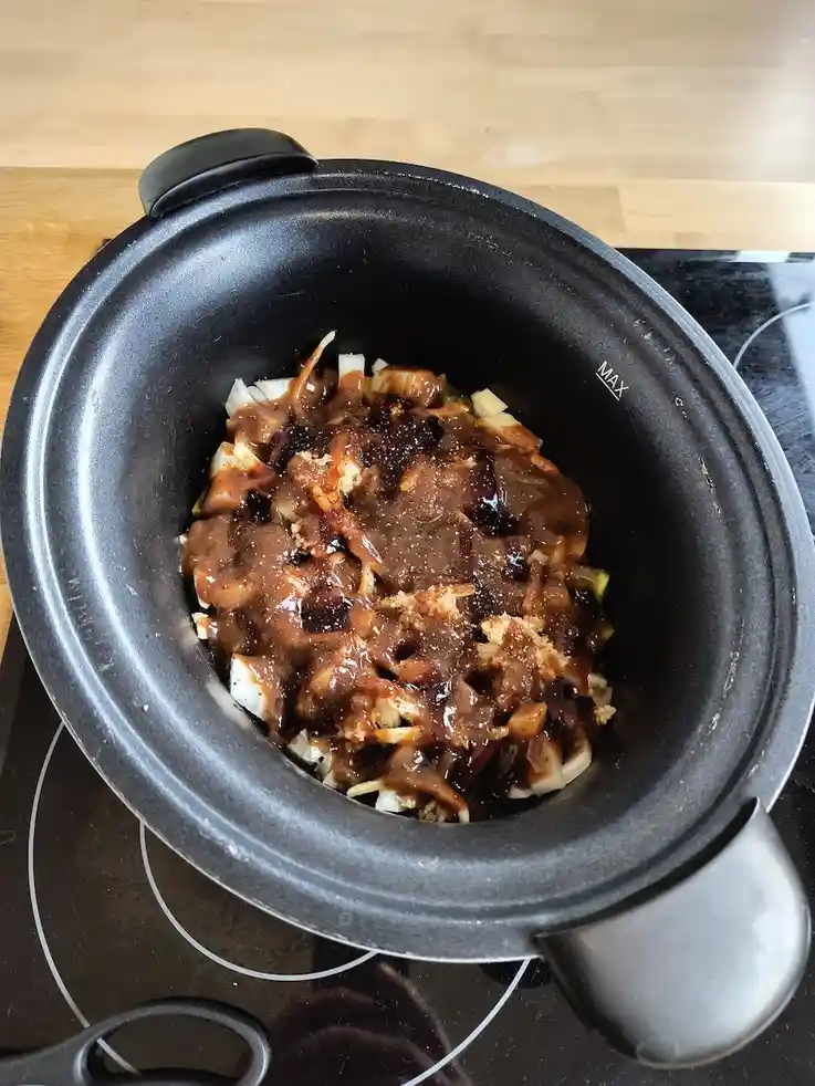 A black slow cooker sits on a black glass stovetop, filled with chopped onions and a thick, dark brown sauce. The text…