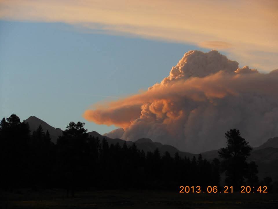 A photograph of a large, billowing smoke cloud, tinged orange by sunset, rising over mountain silhouettes and trees. The…
