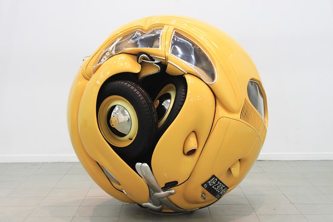 A bright yellow vintage car is transformed into a perfect sphere, with its wheels, windows, and body panels curved to form a…