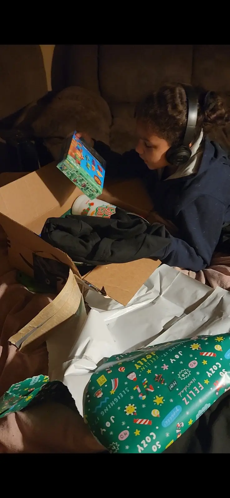 A young person wearing headphones sits on a couch surrounded by unwrapped Christmas presents and a cardboard box, holding up…