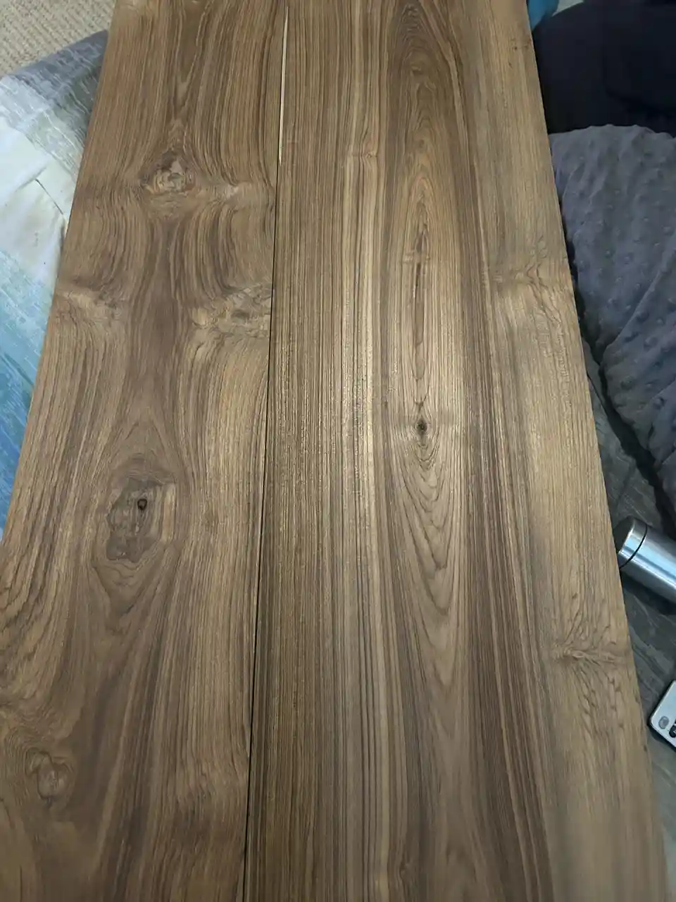 A close-up photo shows a long, light-brown wooden plank with prominent grain patterns and knots, resting on a bed with…