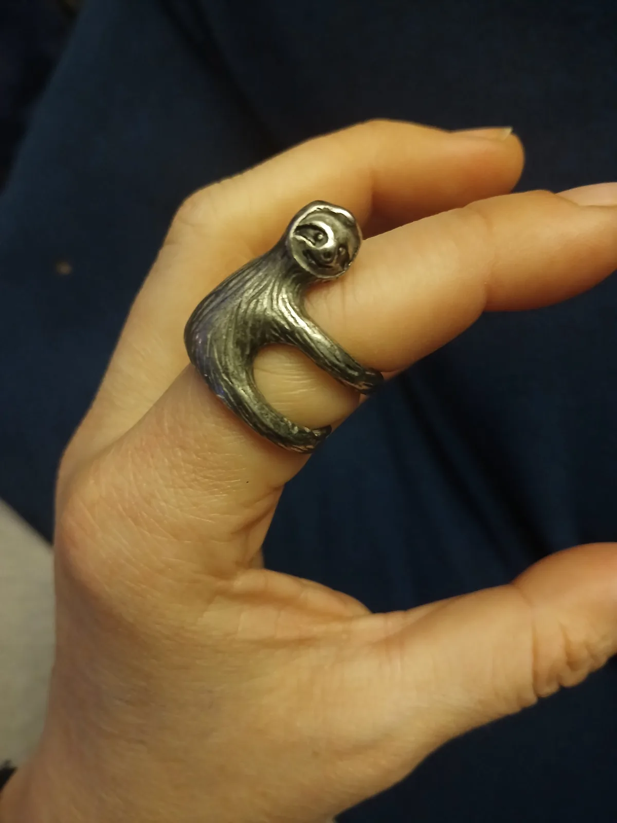 A close-up photo shows a person's hand wearing a large, silver-colored ring shaped like a sloth clinging to the finger. The…
