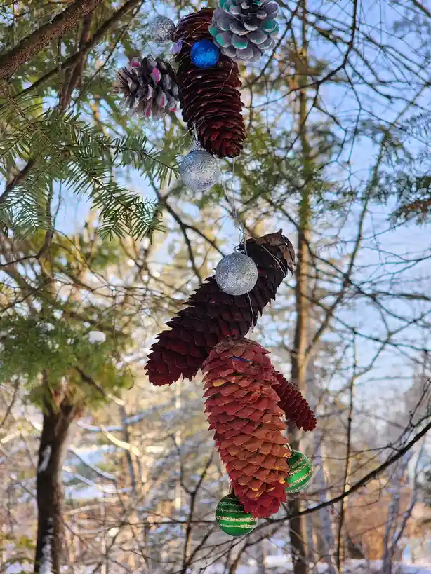 A pinecone is decorated with colorful ornaments and strung with lights, hanging from a tree branch in a snowy forest. The…