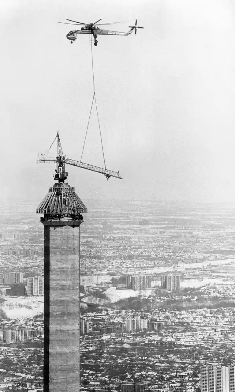 A black-and-white photo shows a helicopter suspended above a tall tower, using cables to lift a large crane assembly that is…