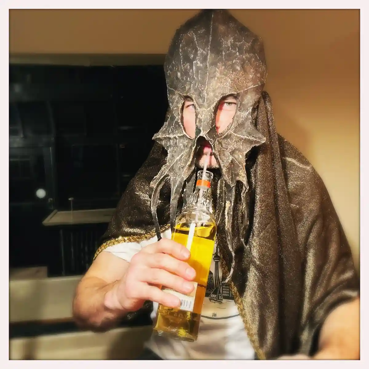 A person wearing a homemade, textured, dark brown kraken costume mask with tentacle-like extensions and a cape, sipping from…