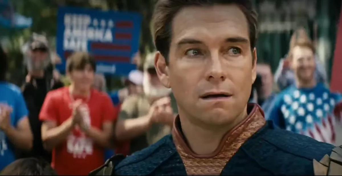 A close-up of a man in a blue and gold costume with a shocked expression, looking upward. Behind him, a blurred crowd…