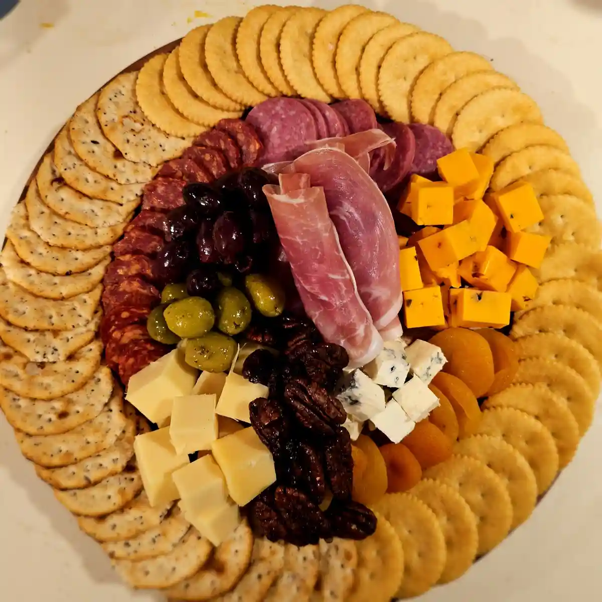A top-down view of a circular charcuterie board filled with neatly arranged crackers, sliced salami, prosciutto, cubes of…