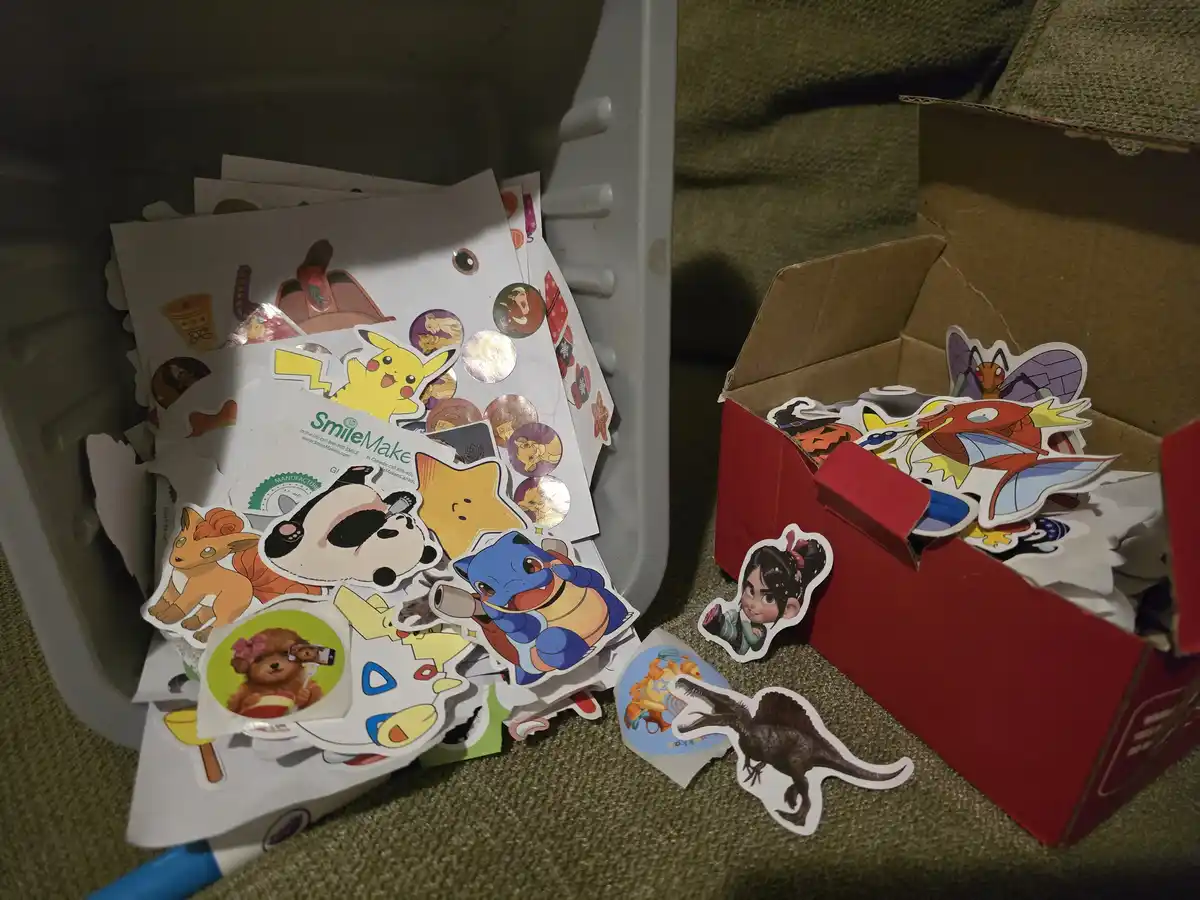 A messy pile of colorful stickers featuring characters like Pikachu, a panda, and a dinosaur spills out of a gray plastic…