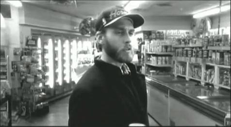 A black-and-white image of a man with a beard and a baseball cap standing in a convenience store aisle, looking slightly…