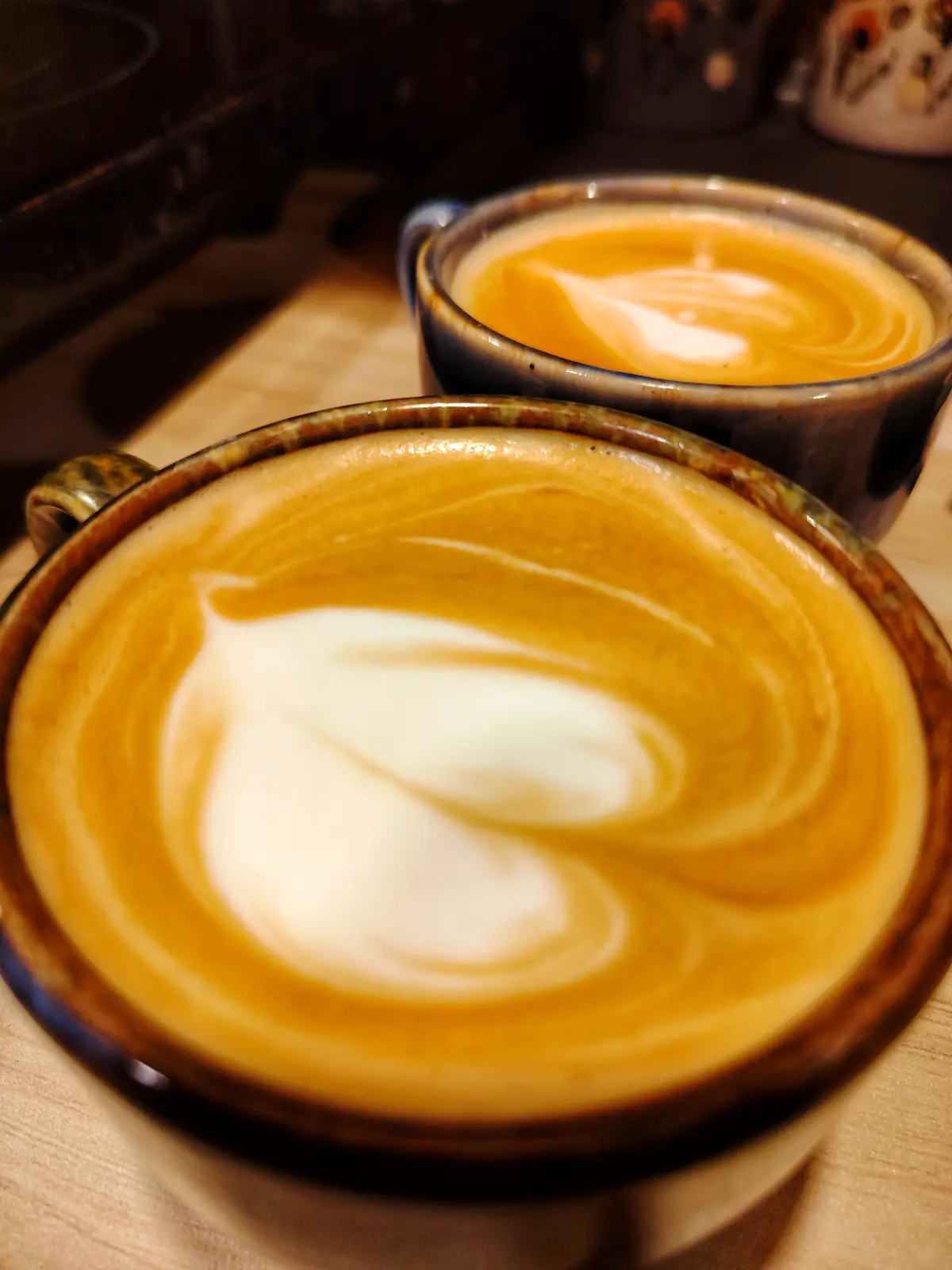 Two ceramic mugs filled with coffee featuring intricate latte art, resting on a wooden surface. The foreground cup has a…