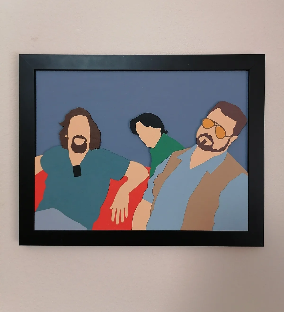 A framed, stylized wooden cutout artwork depicts three figures: a bearded man in a teal shirt on the left, a person with…
