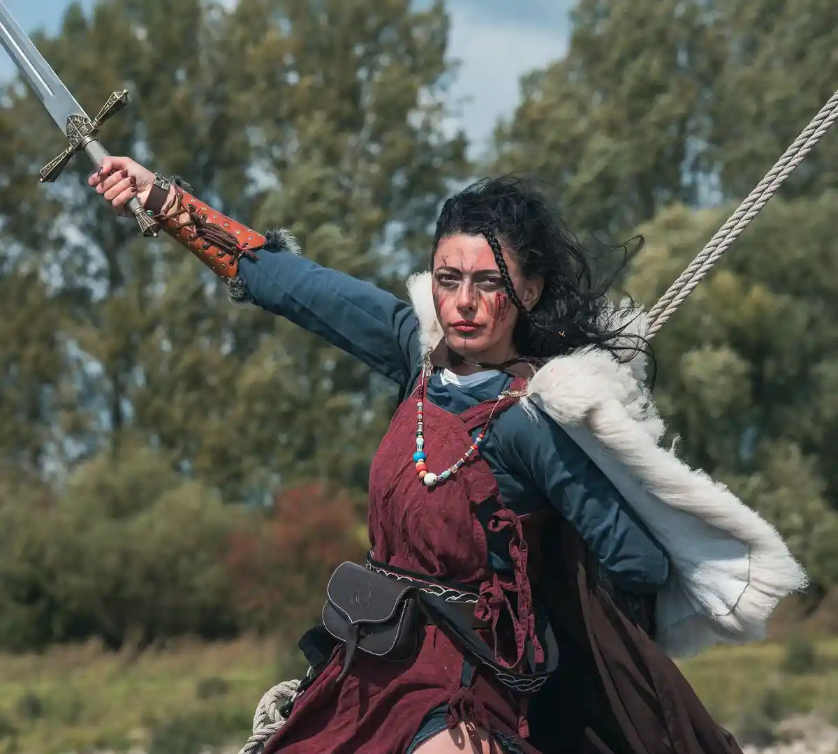 A woman in medieval-style warrior attire, including a fur-lined cloak, leather bracers, and a sword, is suspended by a rope…