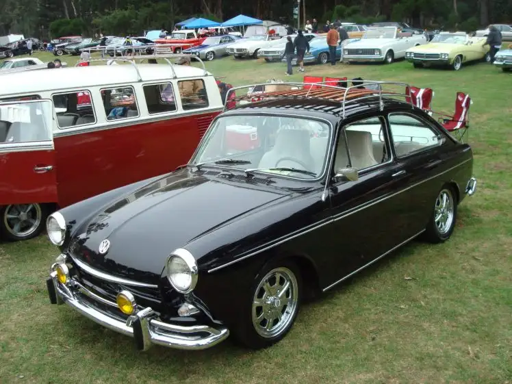A black vintage Volkswagen 1300L with a roof rack is parked on grass at a car show, with a red and white VW bus behind it…