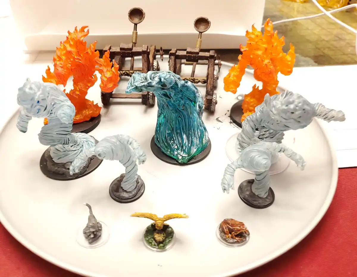 A white plate holds several painted miniature fantasy figures: two large translucent blue/grey humanoid figures, a central…