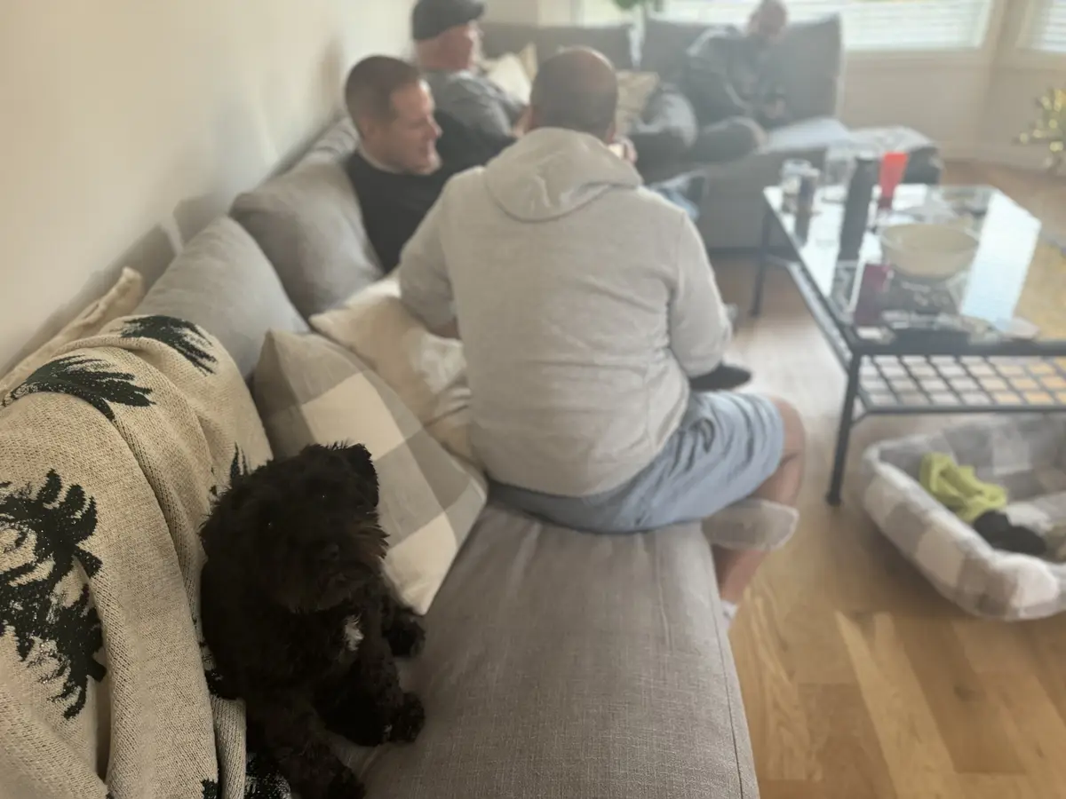 A small black dog sits on a couch in the foreground, looking directly at the camera, while several men are blurred in the…