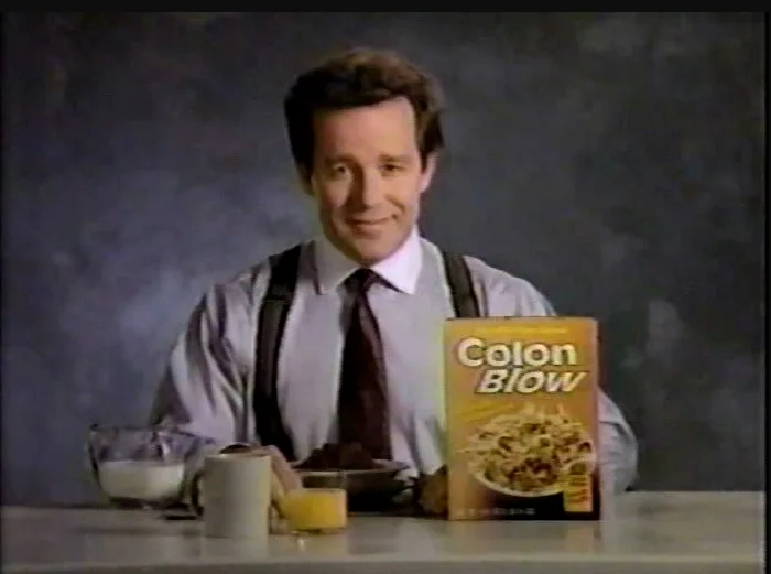 A man in a shirt, tie, and suspenders sits at a table with a bowl of cereal, a glass of milk, and a glass of orange juice…