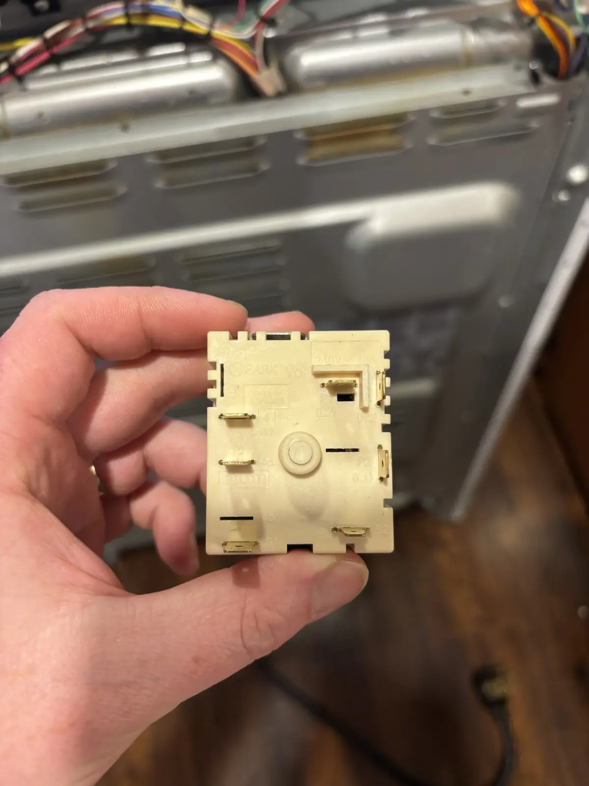 A hand holds a beige, rectangular dryer thermostat control panel with labeled terminals and a central dial, in front of the…
