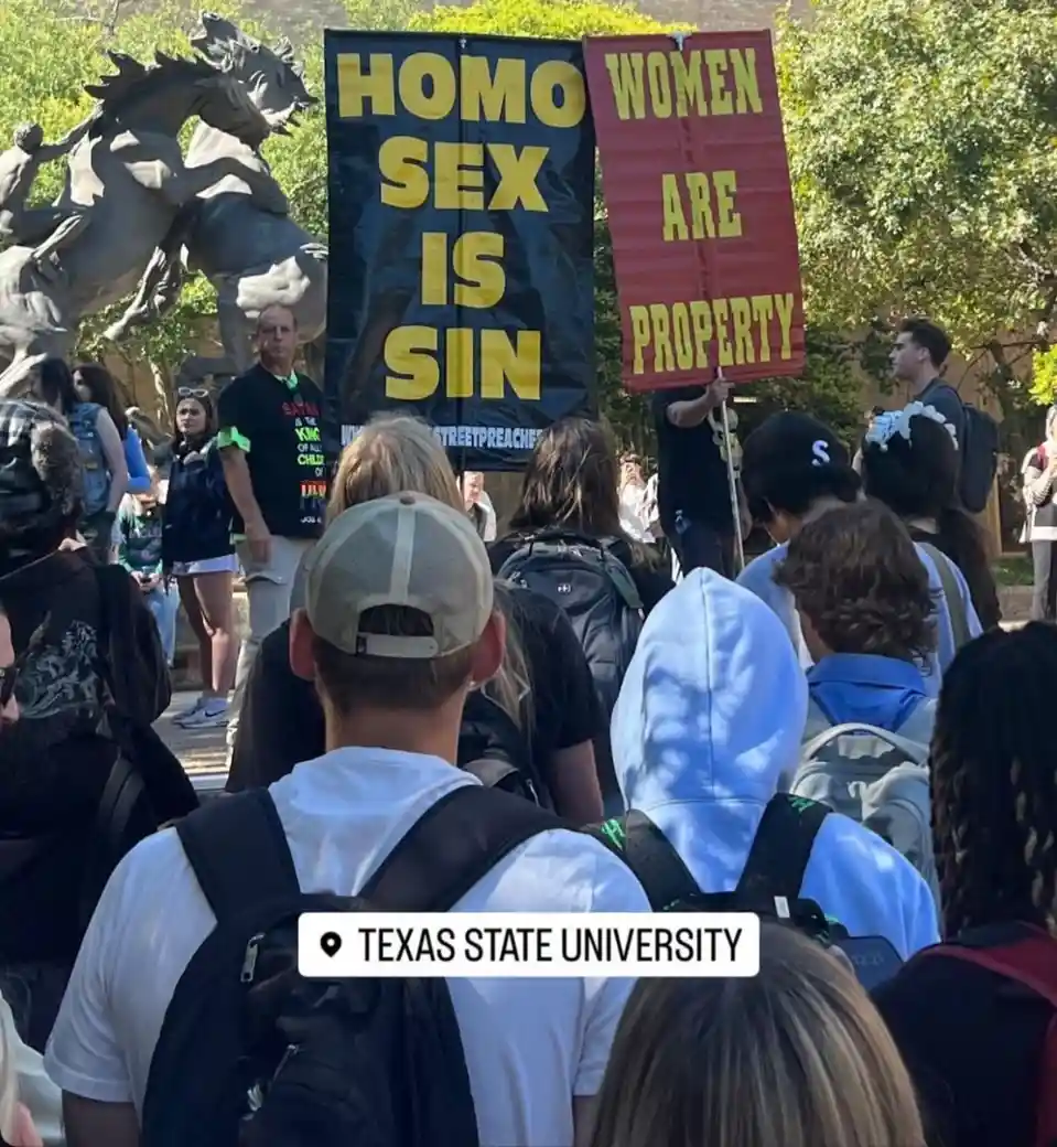 A crowd of students at Texas State University watches as two protesters hold up signs: one black sign with yellow text…