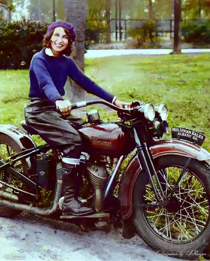 A vintage colorized photograph shows a smiling woman in a purple beret, blue sweater, and leather pants seated on a classic…