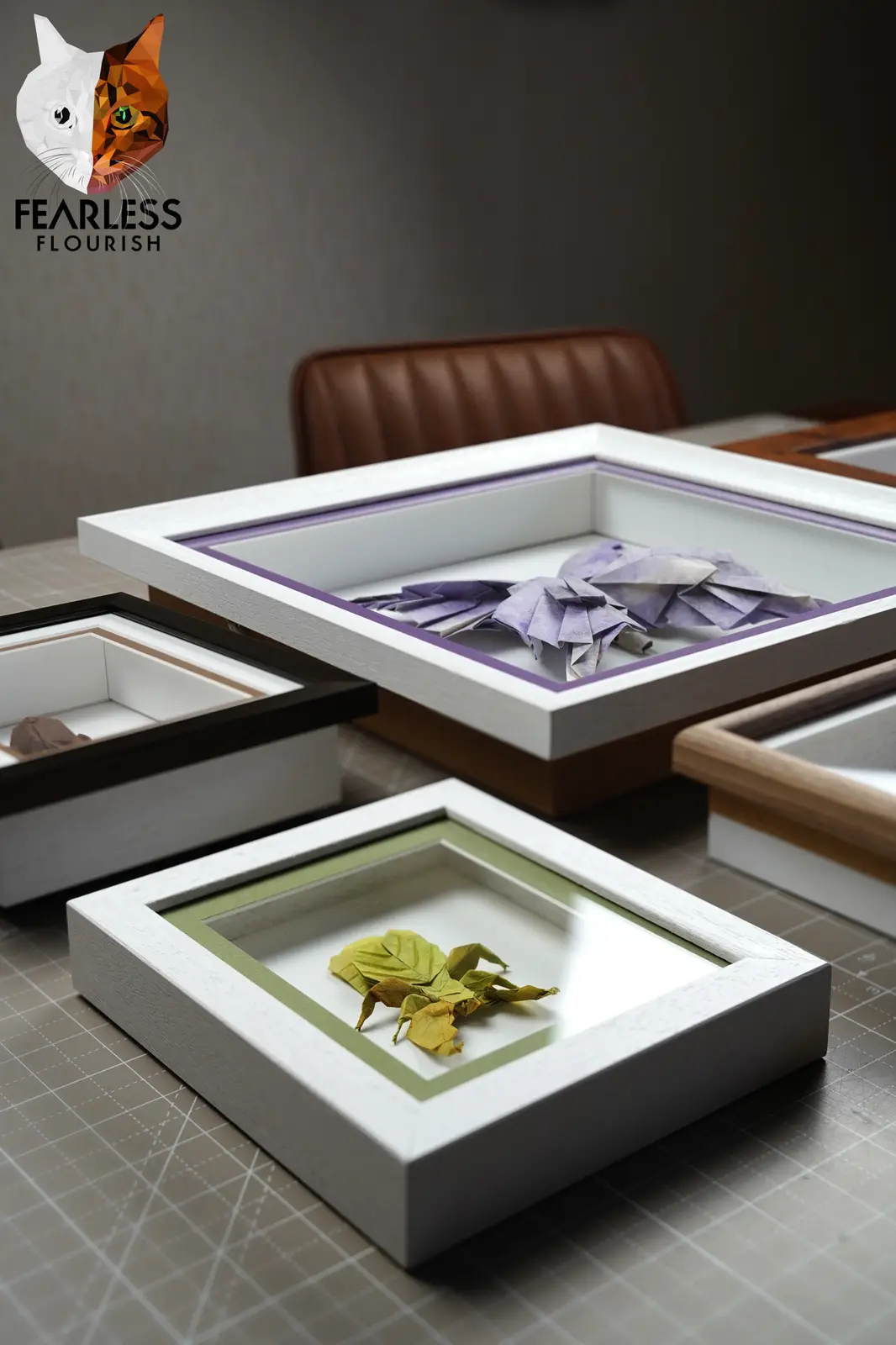 Several framed shadow boxes sit on a gridded work surface, containing dried leaves and purple origami flowers. The top-left…