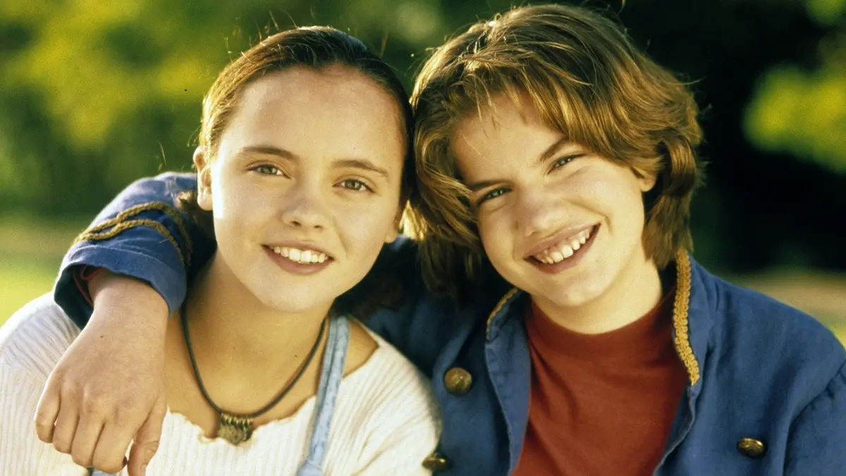 Two young people, a girl with her hair pulled back wearing a white top and a necklace, and a boy with shaggy brown hair in a…
