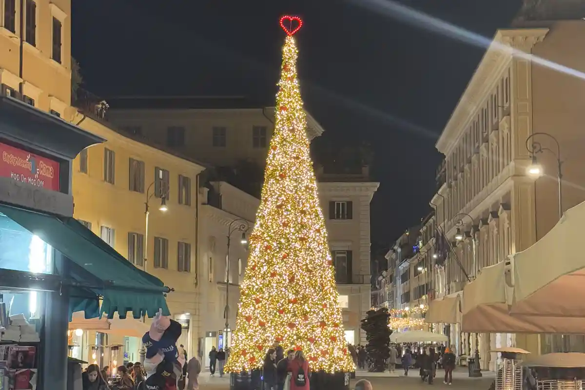 A large, brightly lit Christmas tree stands in a European city square at night, adorned with golden lights and red…