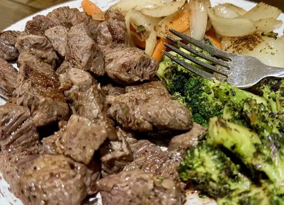 A close-up photo of a plate of food featuring a large pile of cooked, seasoned meat chunks, alongside roasted broccoli…