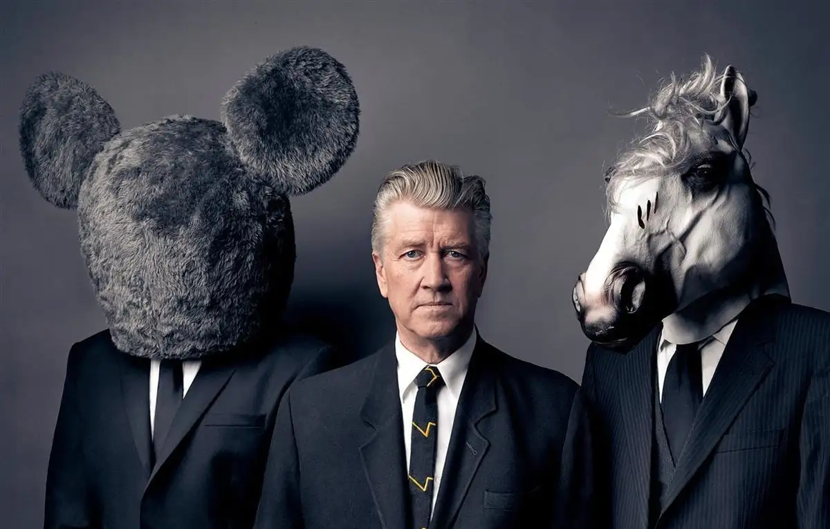 Three men in suits stand against a gray background; the man on the left wears a large gray mouse head, the man on the right…