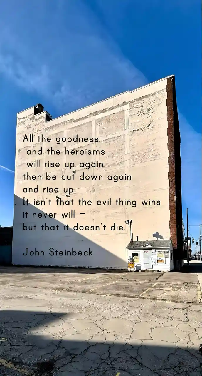 A large, weathered white building wall under a bright blue sky has a quote by John Steinbeck overlaid in black text. The…