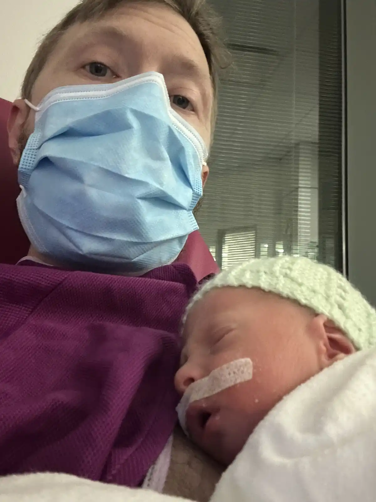 A man wearing a blue surgical mask holds a sleeping newborn baby wrapped in a white blanket. The baby wears a light green…