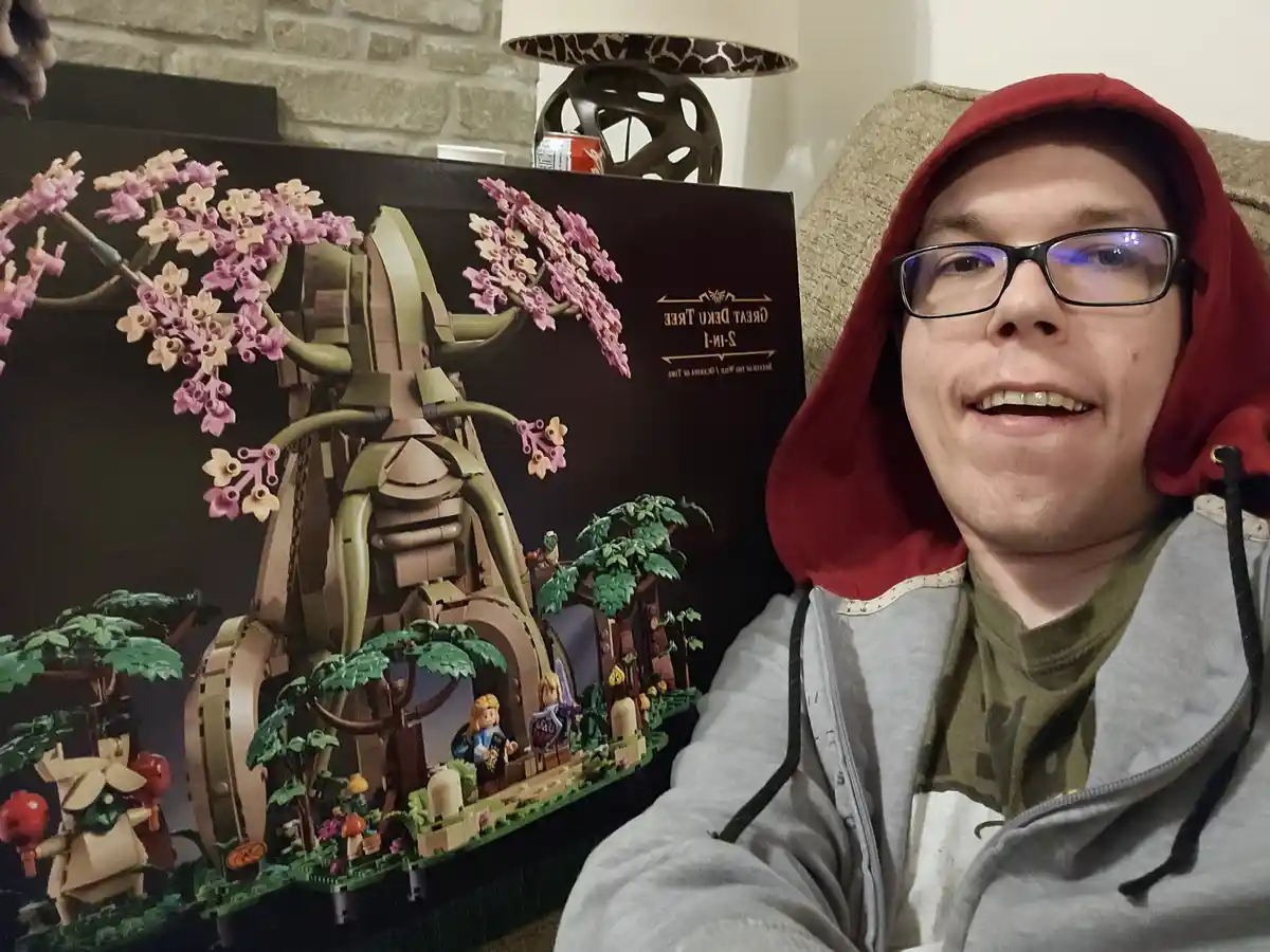 A person in a red hoodie and glasses smiles while sitting beside a large, completed LEGO set of the Great Deku Tree from The…
