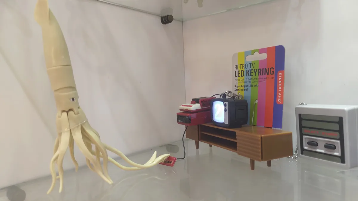 A beige plastic squid toy with one eye and multiple tentacles extends toward a miniature retro TV keyring displaying the…