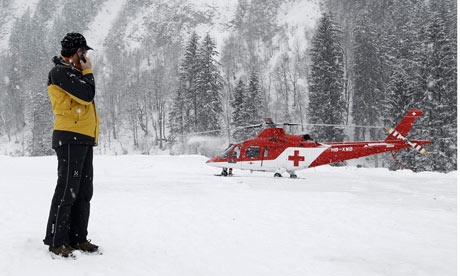 A person in a yellow jacket and black pants stands in a snowy forest, holding a phone to their ear, while a red medical…