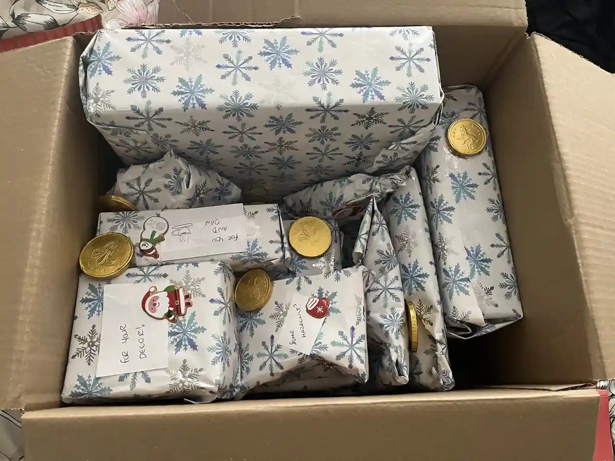 A cardboard box filled with numerous small gifts wrapped in blue and white snowflake-patterned paper. Several gold-colored…