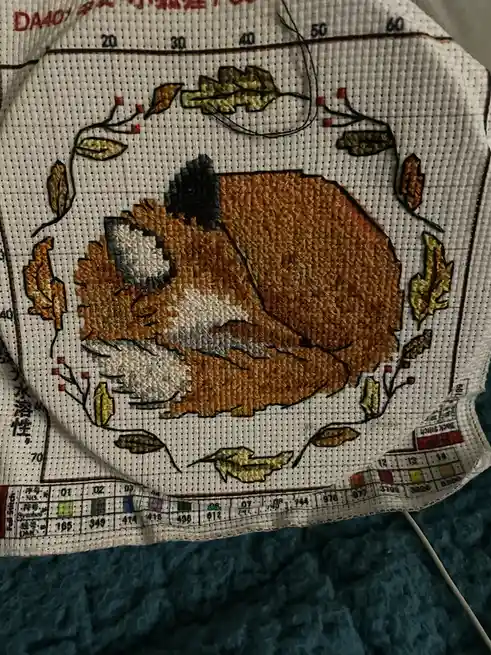 A close-up of a cross-stitch pattern featuring a brown and white cat curled up asleep, surrounded by autumn leaves on a grid…