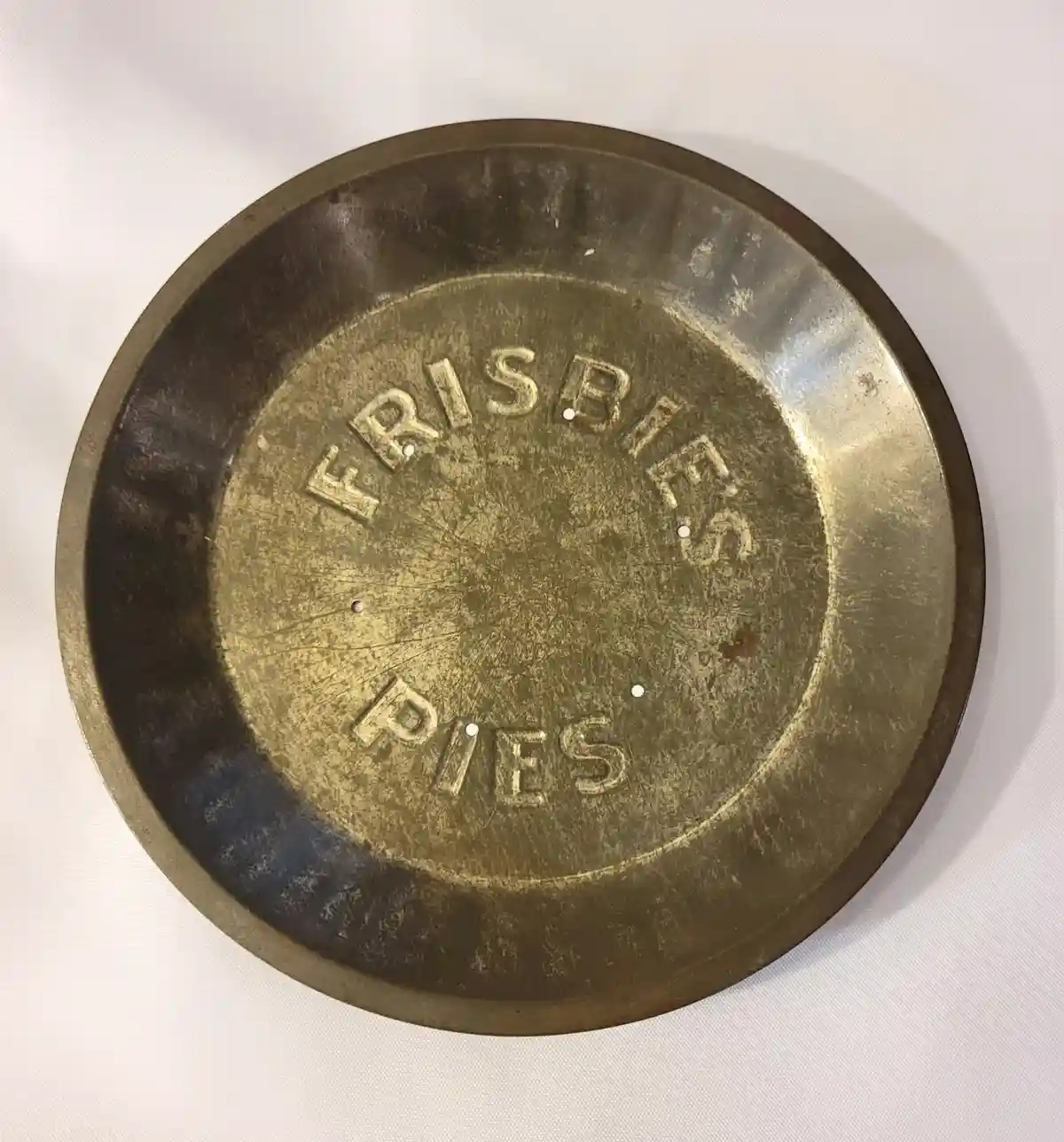 A close-up of a round, tarnished metal pie tin with the words 'FRISBIES' arched above 'PIES' embossed on its surface. The…