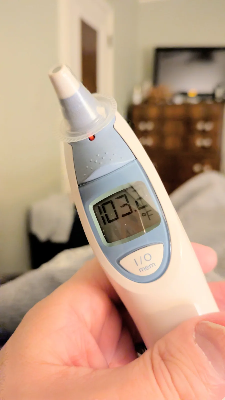 A close-up photo shows a person's hand holding a digital ear thermometer, which displays a high temperature reading of…
