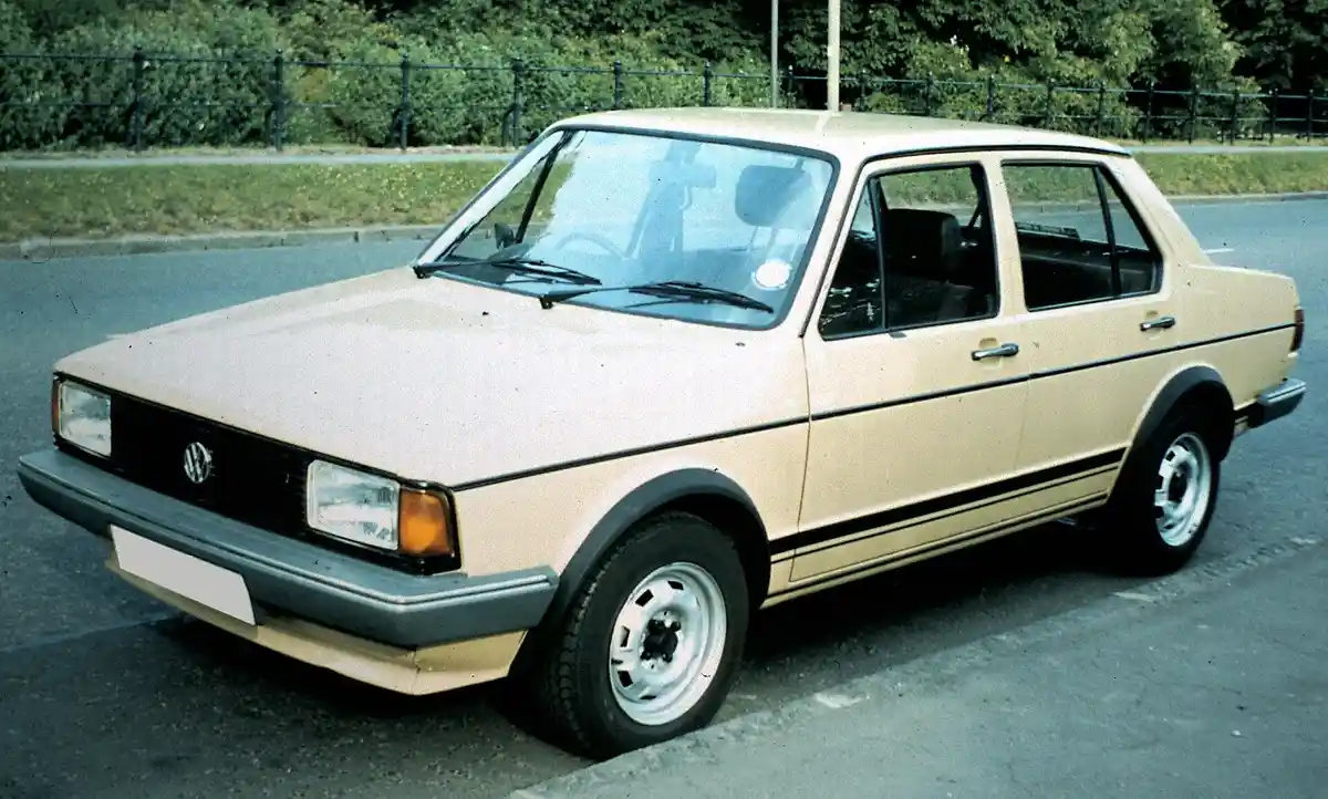 A beige 1980s Volkswagen sedan is parked in an asphalt lot with a black metal fence and green bushes behind it. The car has…