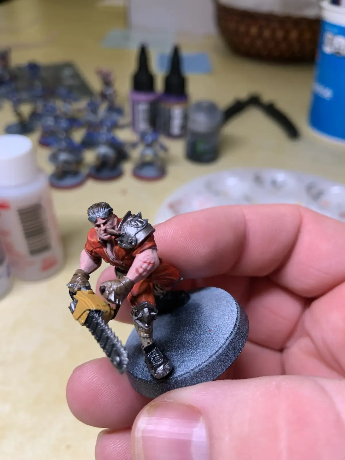 A hand holds a painted miniature of a muscular warrior with a chainsaw, set against a blurred background of other unpainted…